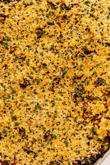 A close-up of a baked casserole topped with a golden, crispy breadcrumb and cheese layer, garnished with chopped herbs in a white dish.