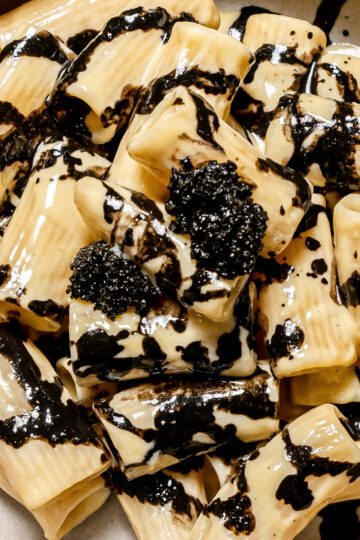 A close-up of rigatoni pasta covered in a creamy sauce with a drizzle and dollops of black squid ink, served on a white plate.
