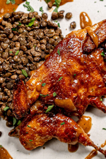 Roasted chicken glazed with sauce, garnished with chopped herbs, served alongside a mound of cooked lentils on a white plate.