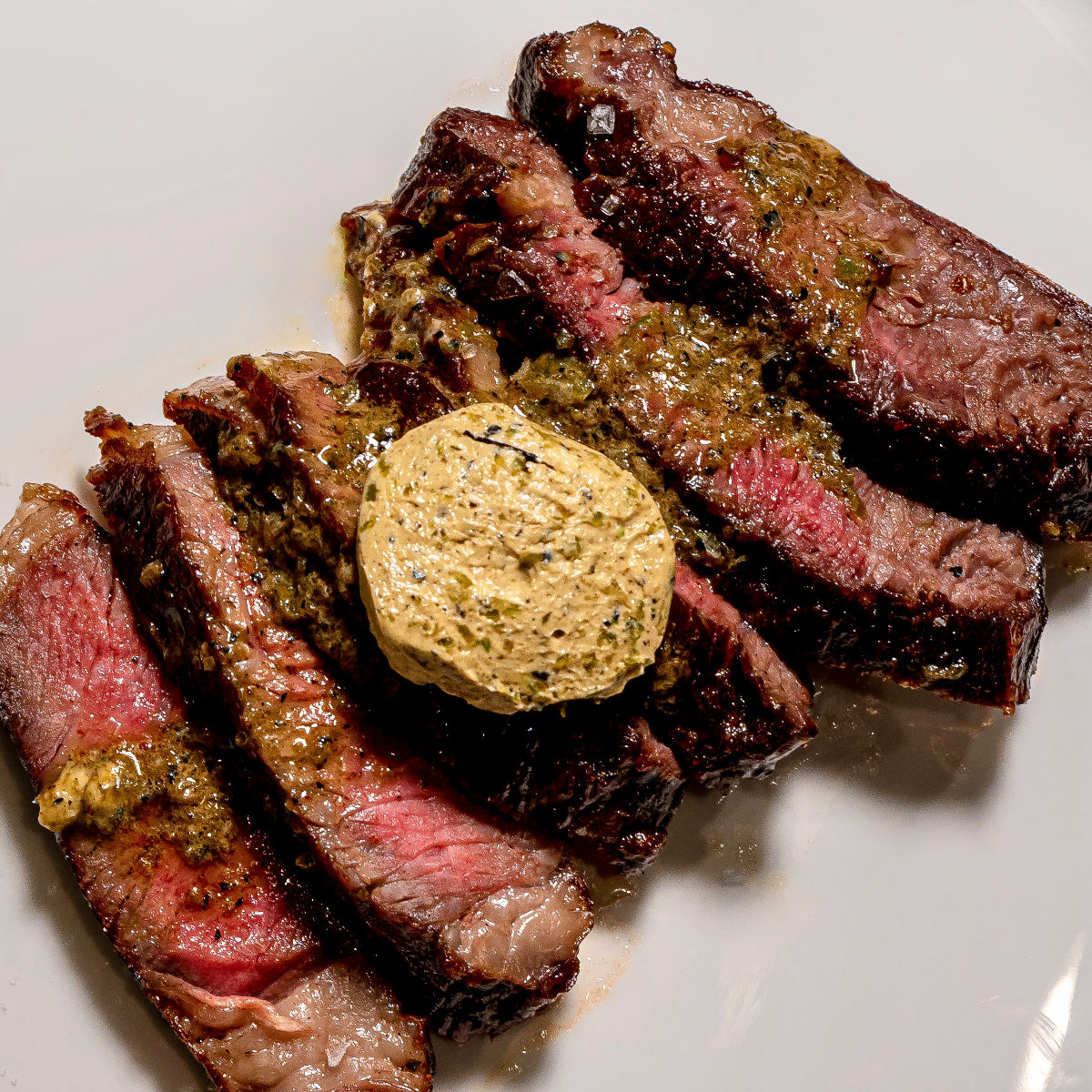 Sliced medium-rare steak topped with a pat of herb butter and drizzled with a green herb sauce, served on a white plate.