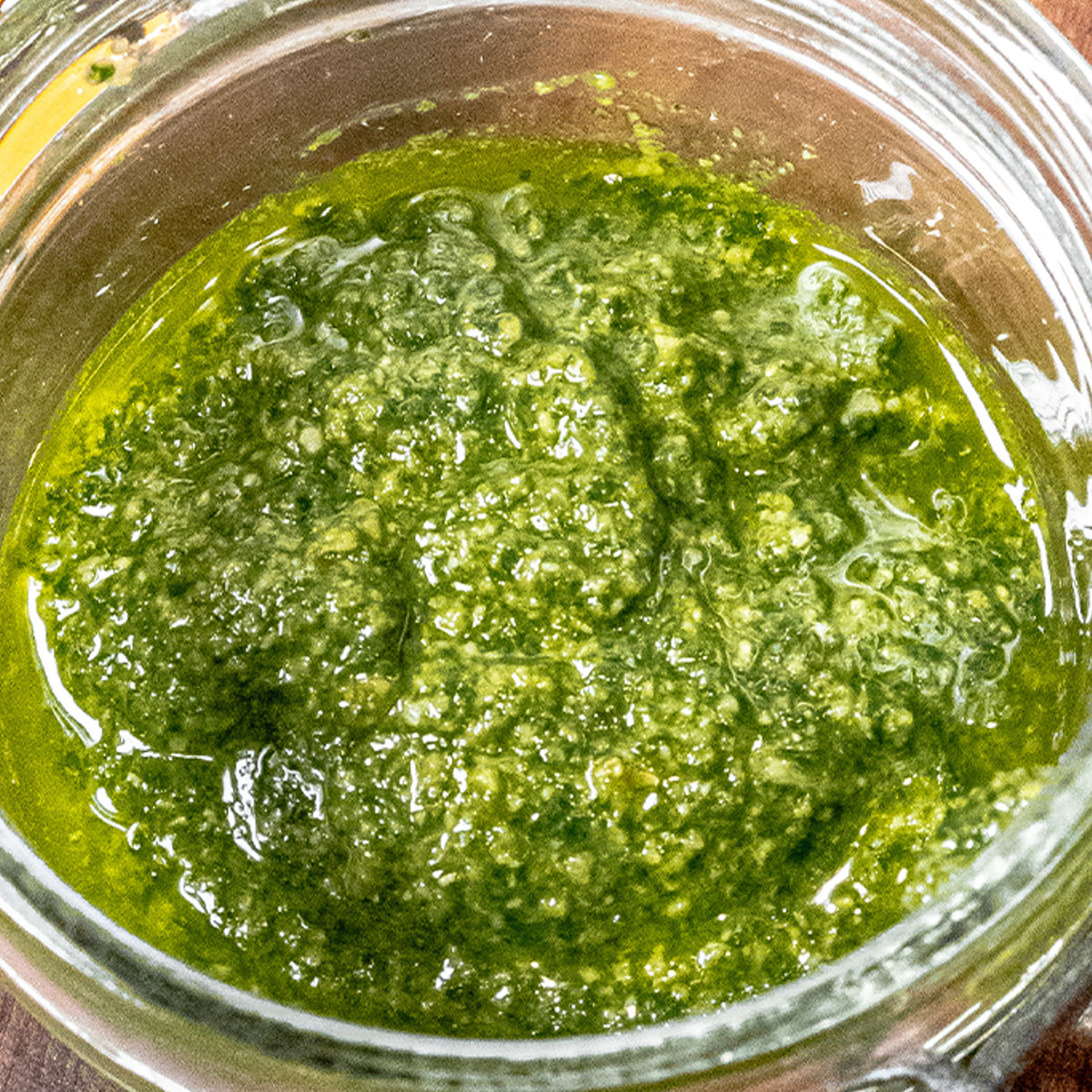 A close-up of a glass jar filled with vibrant green pesto sauce, showing a textured mixture of basil, oil, and other ingredients.