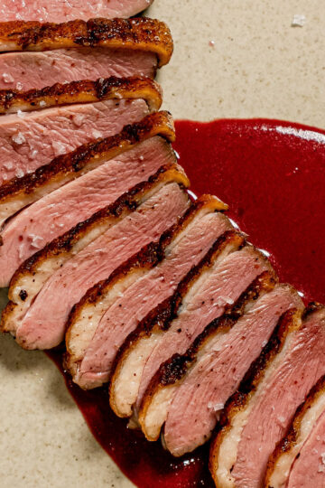 Sliced duck breast with crispy skin arranged in a fan shape on a plate, served with a deep red sauce underneath and sprinkled with coarse salt.