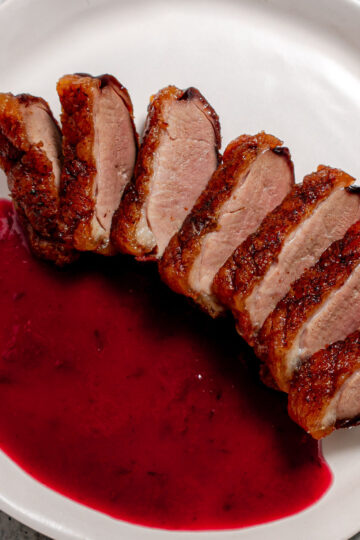 Sliced roasted duck breast with crispy skin arranged on a white plate, served alongside a pool of vibrant red berry sauce.