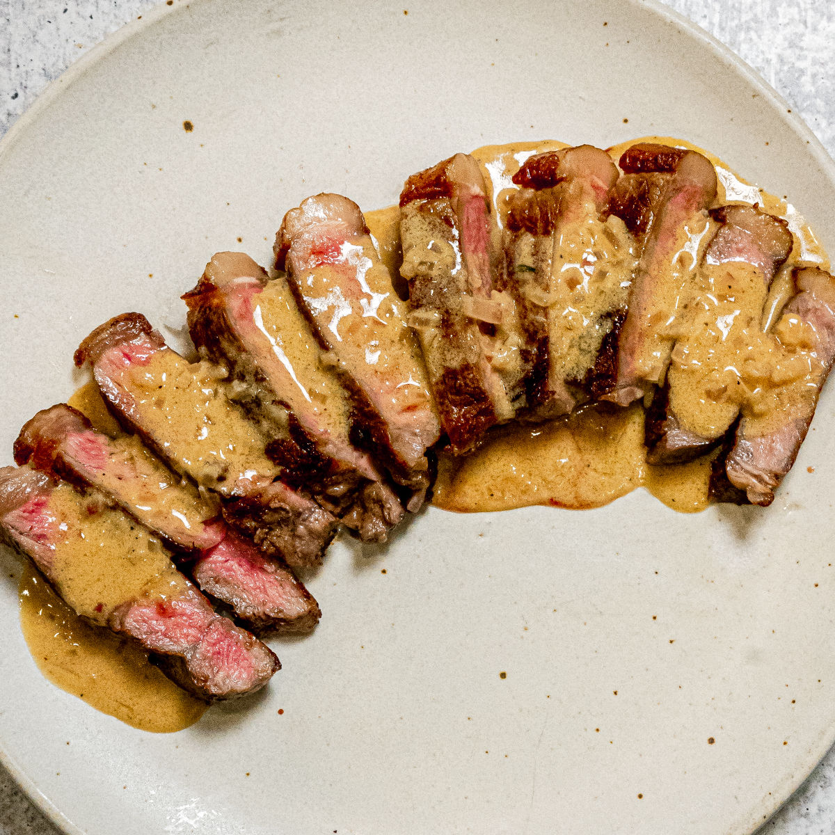 Sliced medium-rare steak arranged on a white plate, topped with a creamy brown sauce. The meat is juicy with a pink center and slightly charred edges.