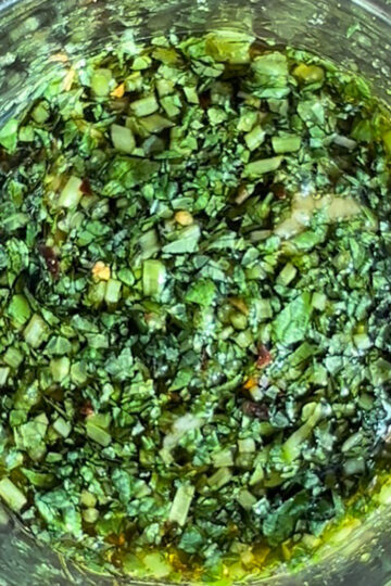 Close-up view of a glass jar filled with finely chopped fresh green herbs mixed with oil, possibly a chimichurri or herb marinade, with the ingredients appearing vibrant and well-blended.