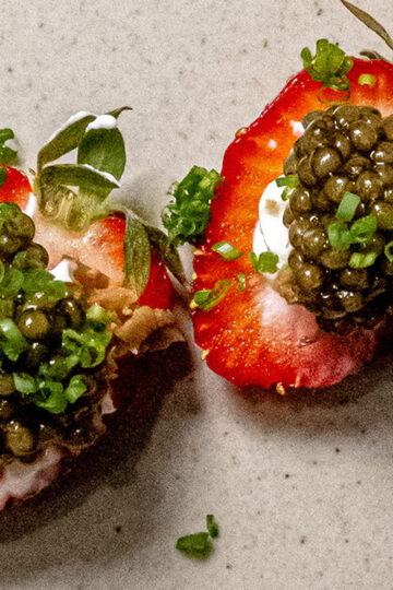 Two strawberry halves topped with a dollop of cream cheese, black caviar, and finely chopped green herbs, placed on a light-colored surface.