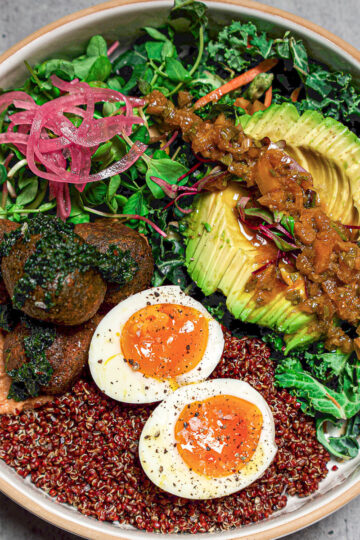 A colorful bowl with quinoa, halved soft-boiled eggs, falafel, sliced avocado topped with salsa, hummus, pickled onions, mixed greens, and herbs, arranged neatly on a gray surface.