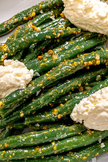 Green beans tossed in a mustard seed dressing, served with three generous dollops of creamy ricotta cheese on a white plate.