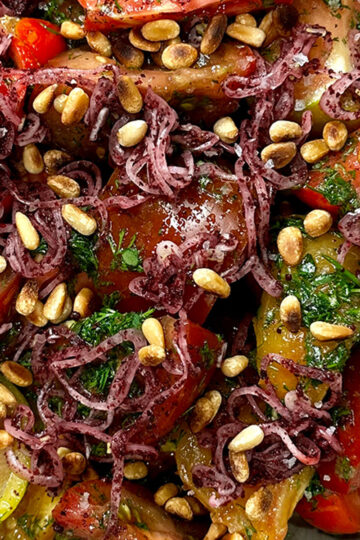 Close-up of a colorful salad with sliced tomatoes, toasted pine nuts, fresh herbs, and thin purple shreds, possibly pickled onions or seaweed, garnished with dressing.