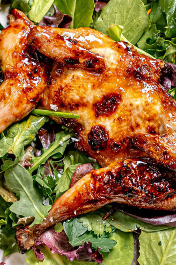 A roasted quail with golden, crispy skin is served on a bed of fresh mixed salad greens, including arugula and spinach, with a glossy glaze on the bird.