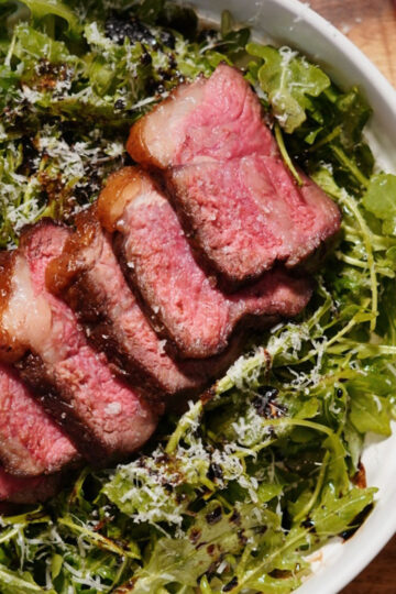 A bowl of fresh arugula salad topped with grated cheese and slices of medium-rare steak, drizzled with balsamic glaze. A red package is partially visible on the wooden table beside the bowl.