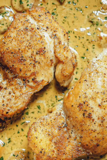 Two pieces of seasoned, pan-seared chicken resting in a creamy, herb-speckled sauce with visible mushroom slices. The chicken has a golden-brown crust and sits in a rich, savory mixture.