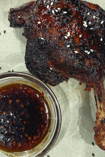 A cooked duck leg with crispy, seasoned skin sits on a plate next to a small glass bowl of dark dipping sauce. Both are sprinkled with black and white sesame seeds.