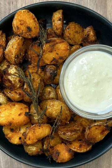 A black plate of crispy roasted baby potatoes garnished with herbs, served with a small bowl of creamy white dipping sauce, on a wooden table.