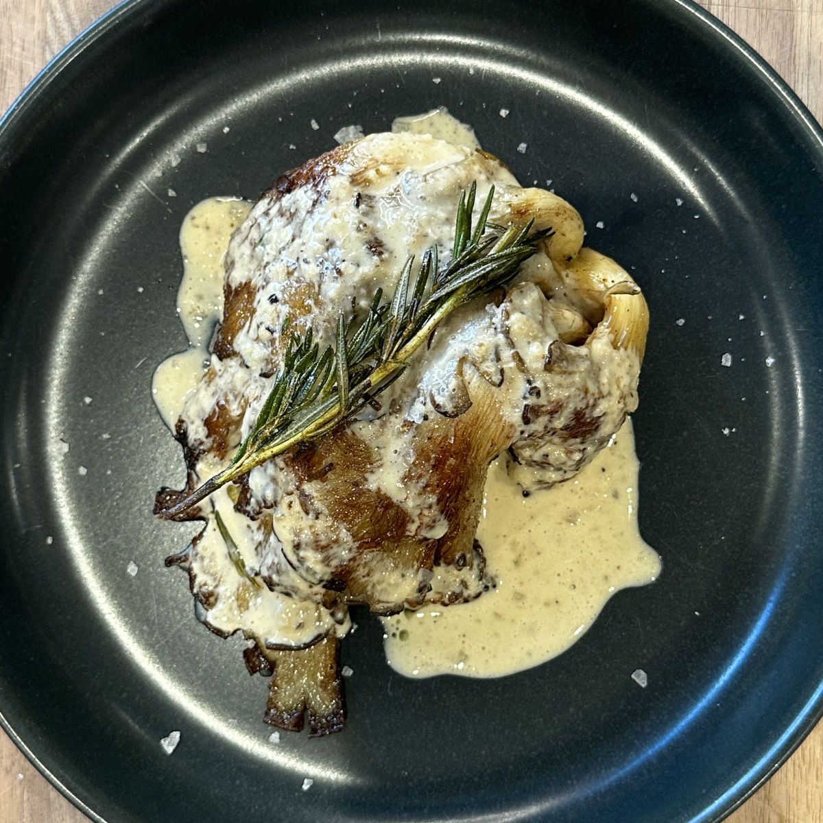 A grilled cauliflower steak topped with creamy sauce and garnished with a sprig of rosemary, served on a dark plate with a sprinkle of coarse salt.