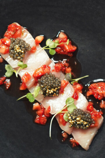 Three pieces of raw fish sashimi topped with black caviar and golden garnish, served with diced red vegetables and microgreens on a dark plate.