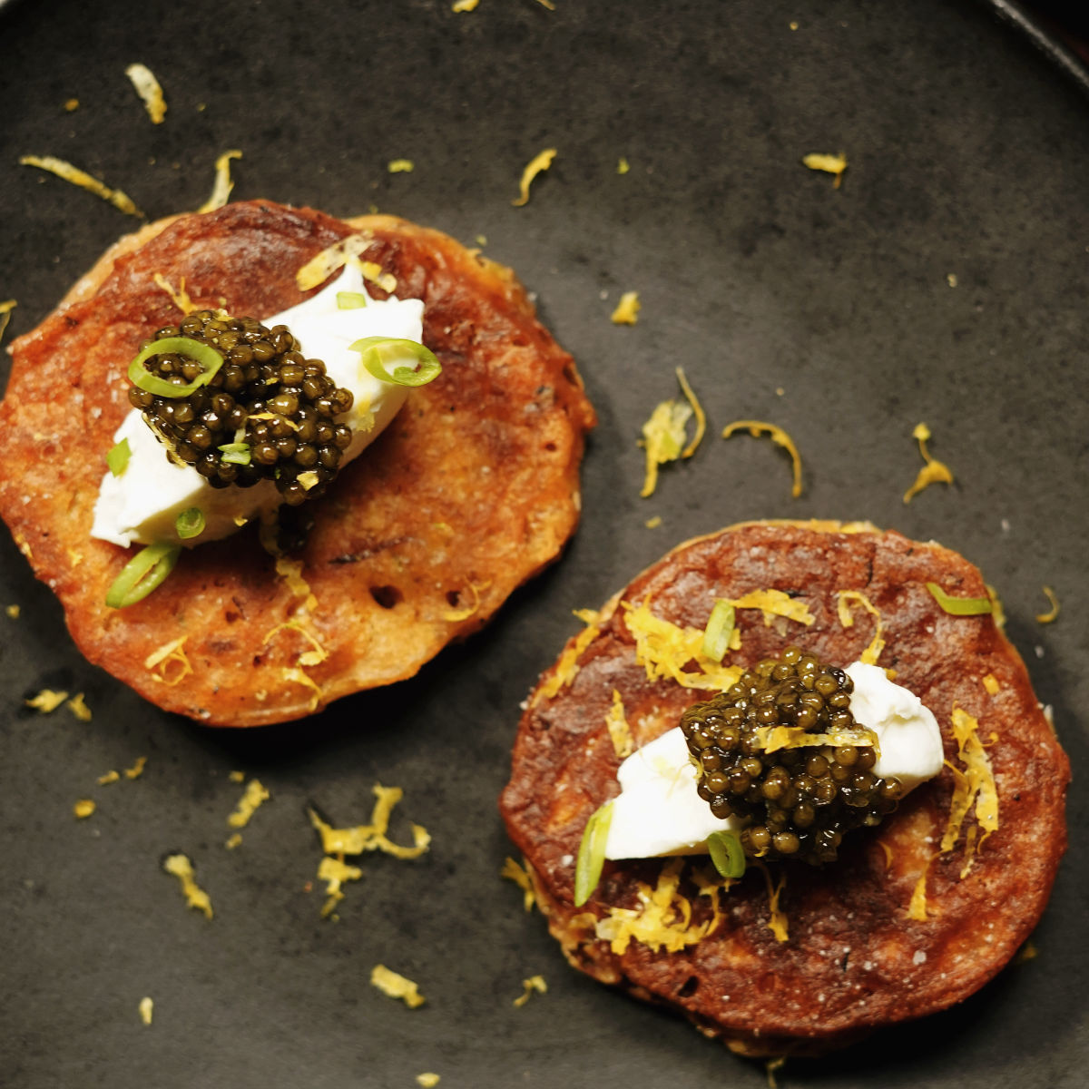 Two blinis topped with dollops of cream, black caviar, chopped green onions, and lemon zest are arranged on a dark plate, with scattered bits of zest around them.