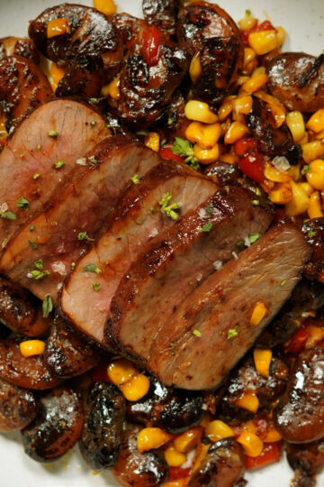 Sliced steak served on a plate with roasted mushrooms and corn, garnished with herbs. The dish appears juicy and well-seasoned, with vibrant colors from the corn and mushrooms.