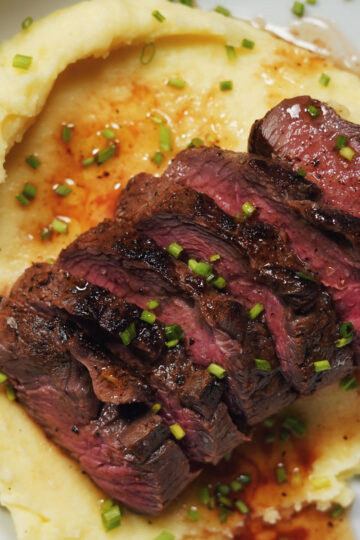 Sliced medium-rare steak served on creamy mashed potatoes, garnished with chopped chives and topped with a drizzle of brown sauce.