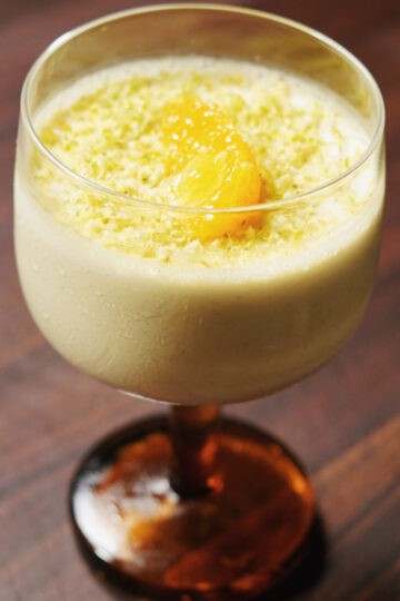 A creamy dessert served in a glass with an orange base, topped with a dollop of orange fruit and grated zest, placed on a wooden surface.