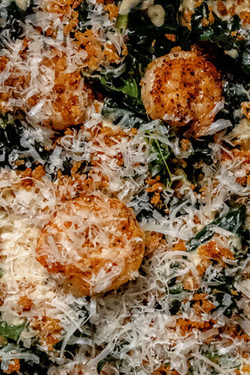 Close-up of a dish featuring seared scallops topped with grated cheese, crispy breadcrumbs, and wilted greens, all seasoned and served together in a savory presentation.