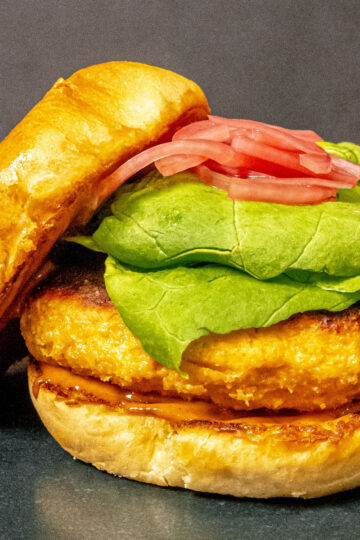 A close-up of a sandwich with a golden, crispy patty, fresh green lettuce, pickled red onions, and a toasted bun on a dark background.