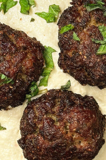 Three browned meatballs sit on a layer of creamy white sauce, garnished with chopped fresh herbs.