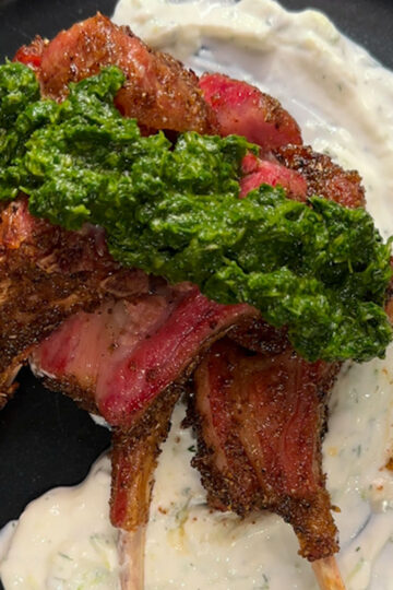 Three cooked lamb chops with a green herb sauce rest on a bed of creamy white yogurt sauce, served on a dark plate. The lamb is well-seasoned, charred, and slightly pink inside.