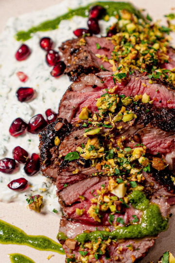 Sliced grilled steak topped with chopped pistachios, served with green sauce, creamy yogurt with herbs, and pomegranate seeds on a light-colored plate.