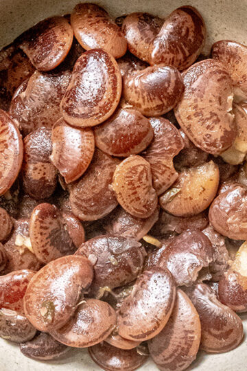 A close-up of a bowl filled with shiny, cooked brown beans that appear to be large and oval-shaped, possibly lima or butter beans, resting in a light-colored ceramic dish.