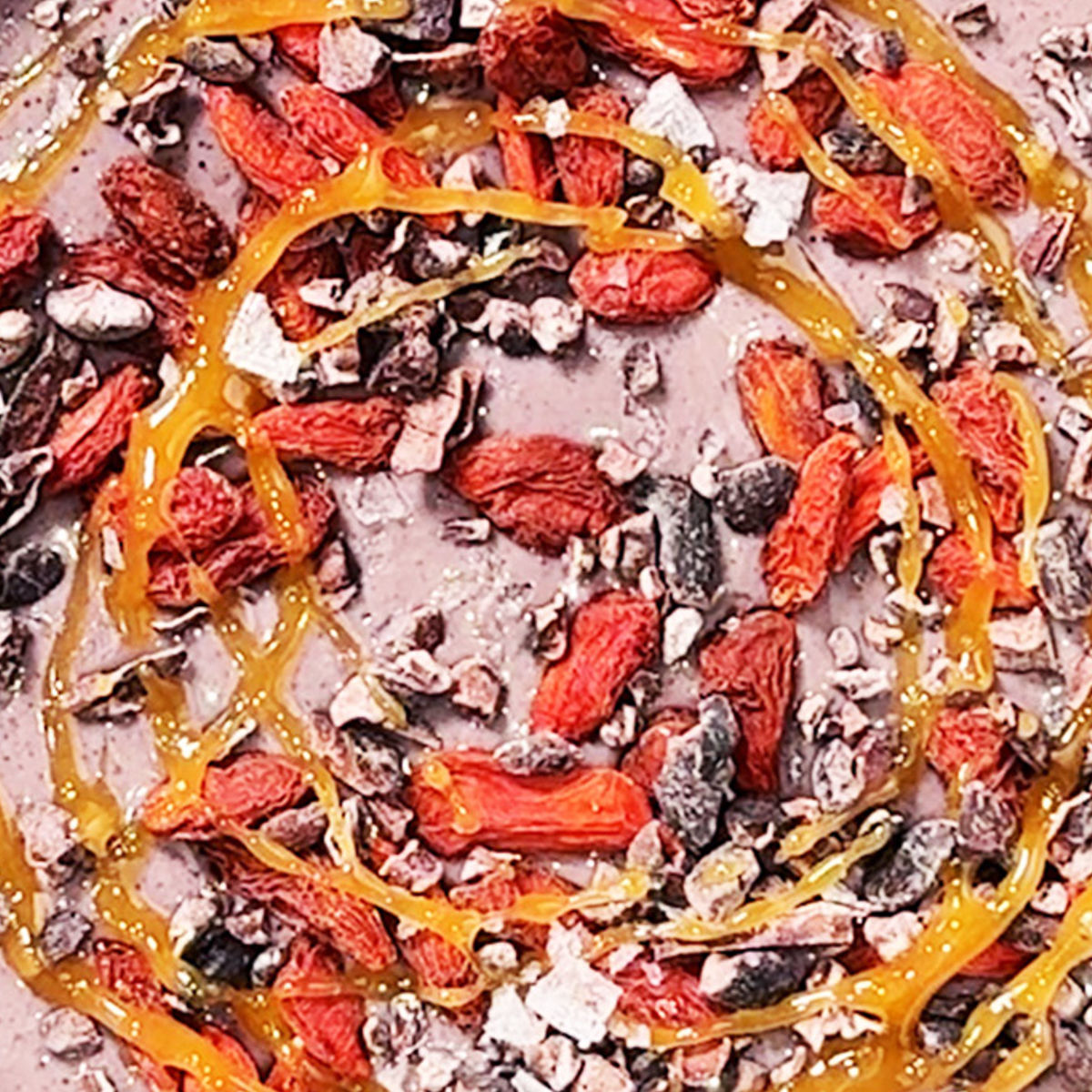 Close-up of a colorful food surface topped with dried goji berries, cacao nibs, and drizzled with golden syrup in a spiral pattern. The background appears creamy and purple-hued.