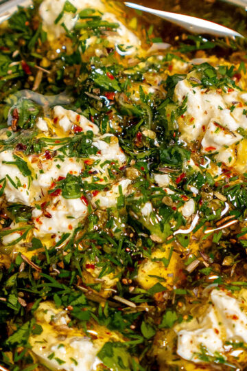Close-up of soft cheese cubes marinated in olive oil with chopped herbs, red pepper flakes, and seasonings in a glass jar. The mixture looks fresh and flavorful.