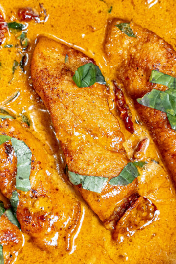 Three pieces of chicken breast in a creamy orange sauce, garnished with chopped fresh basil leaves. The sauce appears rich and spiced, coating the chicken evenly.