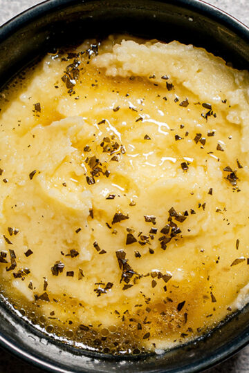 A bowl of creamy mashed potatoes topped with melted butter and sprinkled with dried herbs, seen from above.