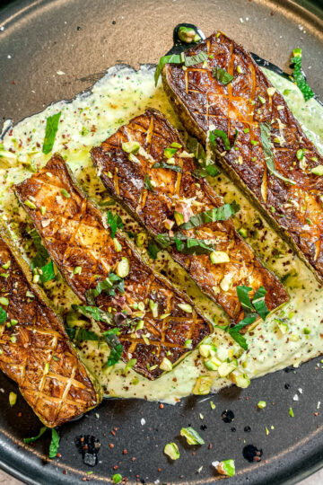 Four pieces of roasted, cross-hatched eggplant are served on a black plate over a creamy green sauce, garnished with chopped herbs and nuts.