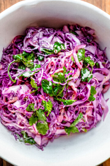 A bowl of colorful coleslaw made with shredded purple cabbage, cilantro, and carrots, displayed on a wooden surface.