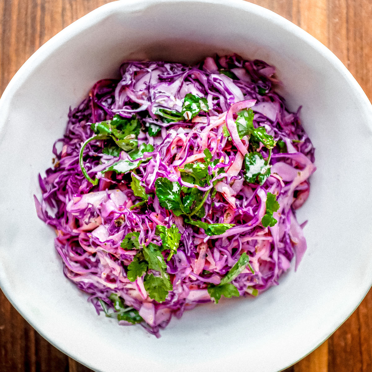 A bowl of colorful coleslaw made with shredded purple cabbage, cilantro, and carrots, displayed on a wooden surface.