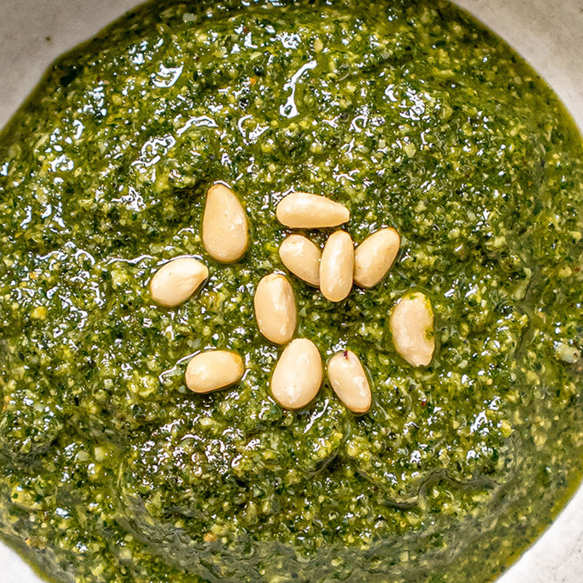 A close-up of green pesto sauce topped with several pine nuts, served in a white bowl.