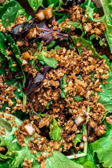 A bowl of mixed green salad topped with crunchy brown breadcrumbs and diced onions, drizzled with a dark vinaigrette dressing.