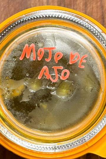 A glass jar with a metal clasp, labeled Mojo de Ajo in red marker on the lid, sits on a wooden surface. The contents inside the jar appear oily with visible herbs or garlic cloves.