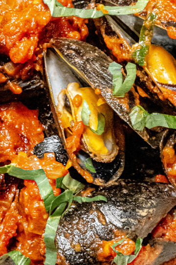 Close-up of cooked mussels in their shells, covered in a vibrant red tomato sauce and garnished with fresh basil leaves.