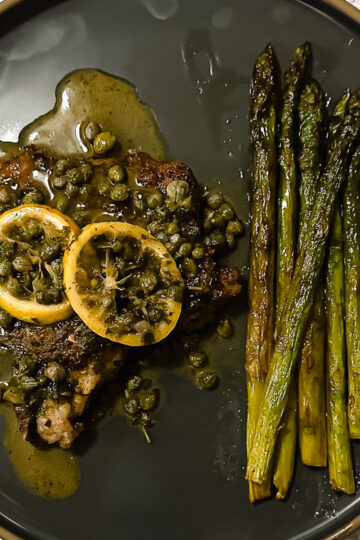 A black plate holding a piece of meat topped with lemon slices, capers, and sauce, served alongside a row of roasted asparagus spears.