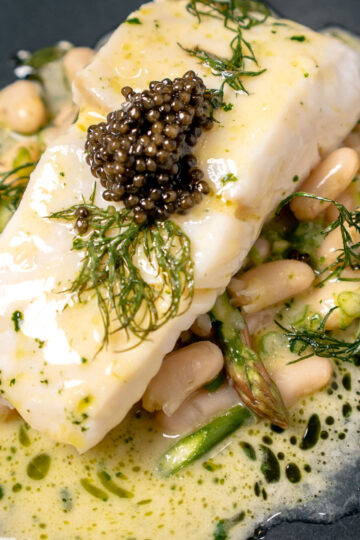 A plate of poached fish topped with caviar and fresh dill, served over white beans and asparagus in a creamy herb sauce.