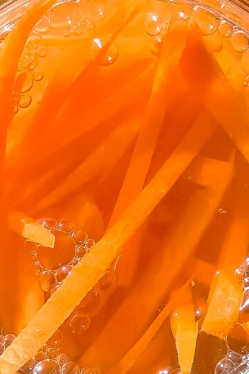 Top view of thin orange carrot sticks submerged in liquid with visible bubbles, likely in a glass jar for pickling or fermenting.