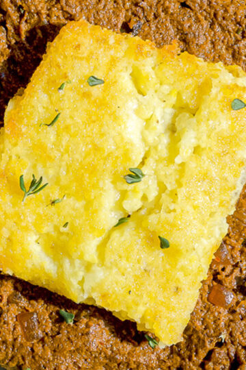 A close-up of a square piece of golden cornbread topped with small green herb leaves, sitting on top of a bed of brown, chunky chili.