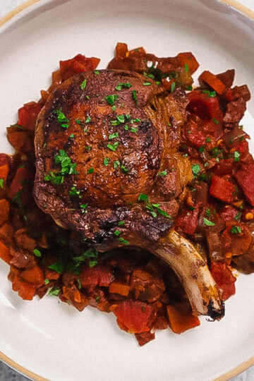 A cooked pork chop sits atop a bed of diced tomatoes, carrots, and other vegetables, garnished with chopped parsley, on a white plate.