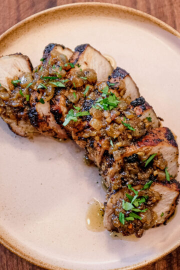 Sliced grilled pork tenderloin topped with a chunky green sauce and garnished with chopped herbs, served on a beige plate atop a wooden surface.