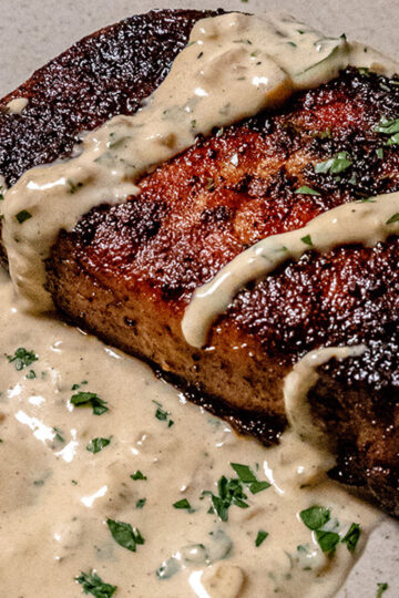 A close-up of a seared steak topped with creamy peppercorn sauce and garnished with chopped parsley, served on a light-colored plate.