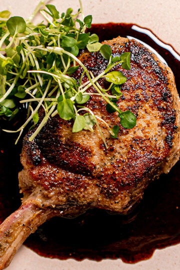 A grilled bone-in pork chop served on a light plate, topped with fresh microgreens and plated over a rich, dark sauce.