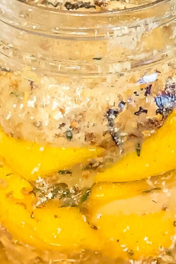 A glass jar filled with lemon slices, herbs, and coarse salt, showing a preserved lemon mixture with visible spices packed between the lemon layers.